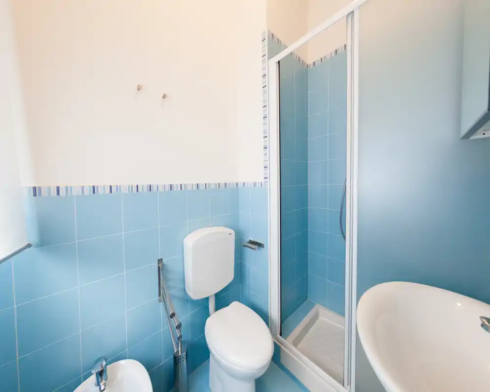 Bagno luminoso e pulito con rivestimenti blu, cabina doccia, wc, bidet e lavabo bianco — ideale per un affitto compatto.