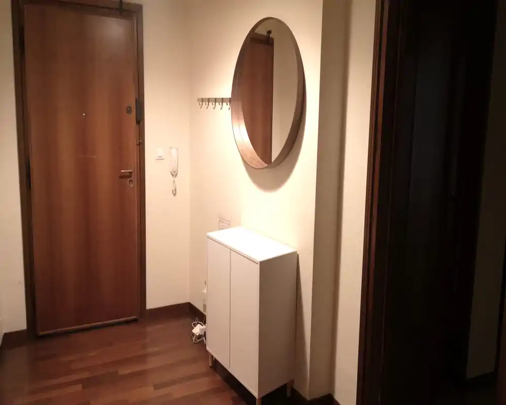 Piccolo ingresso con porta d'ingresso in legno, grande specchio rotondo e mobiletto bianco compatto — illuminazione calda e ordine, perfetto per mostrare l'entrata dell'appartamento.
