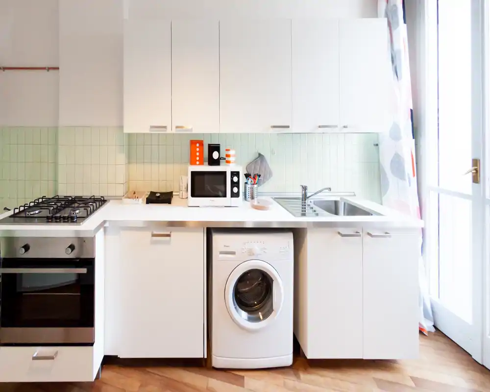 Cucina moderna e luminosa con forno incassato, piano cottura a gas, lavello e lavatrice sotto il piano; mobili bianchi e piastrelle verde chiaro per un ambiente funzionale e pulito.