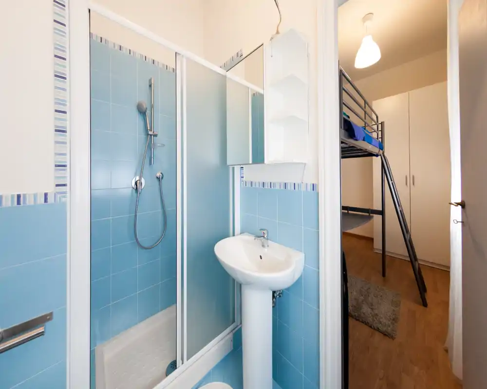 Bagno compatto con doccia piastrellata blu, lavabo a colonna e mobiletto con specchio; ambiente luminoso, pulito e ben illuminato adiacente a una camera con letto a castello.
