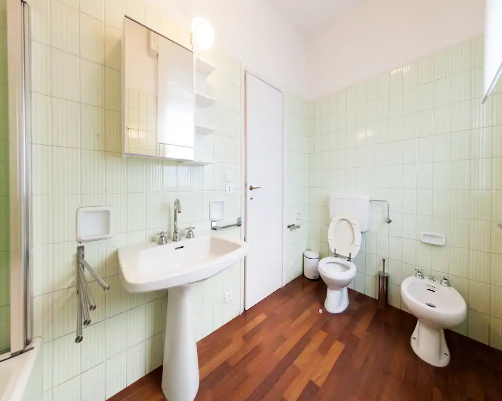 Bagno luminoso e spazioso con lavabo a colonna, water e bidet su pavimento effetto legno e pareti piastrellate verde chiaro, pulito e arioso.