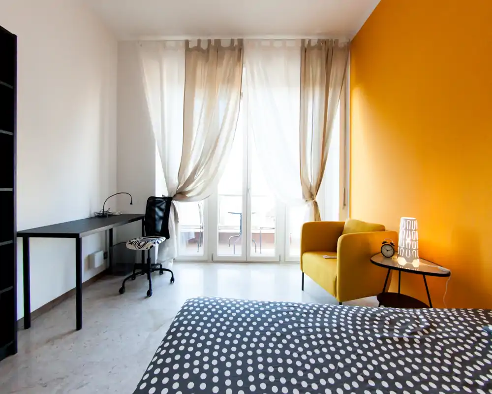 Bright, styled bedroom with a polka-dot bed in the foreground, a yellow accent armchair by the window and a workspace desk. Large glass doors with sheer curtains flood the room with natural light and open to a small balcony.