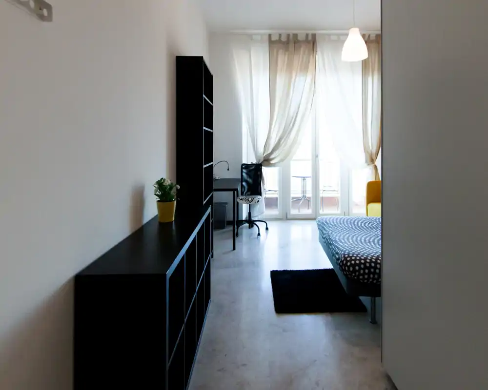 Bright, minimalist bedroom with a patterned bed, desk and balcony access. Natural light fills the room through sheer curtains, creating a calm workspace and sleeping area.