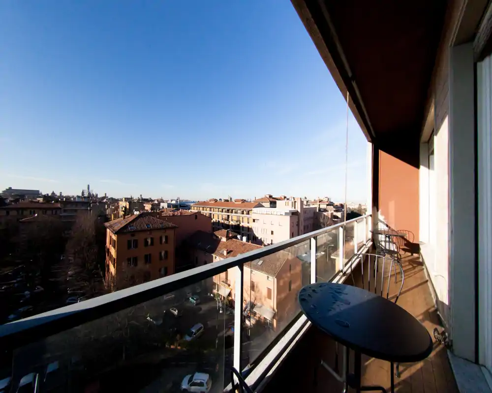 Sunny balcony with glass railing offering a wide city view; small outdoor table and chairs for relaxing and enjoying the panorama.