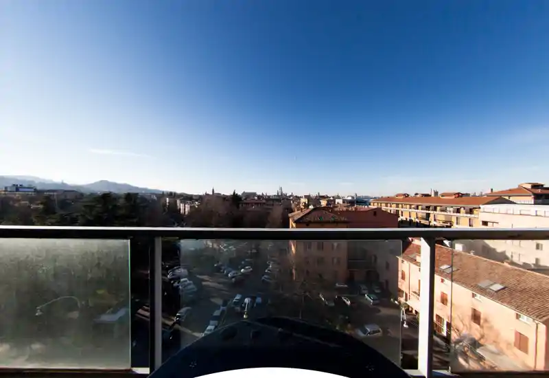 Balcone luminoso con vista sui tetti della città e sulle colline in lontananza, ringhiera in vetro e cielo aperto — ideale per la colazione mattutina.