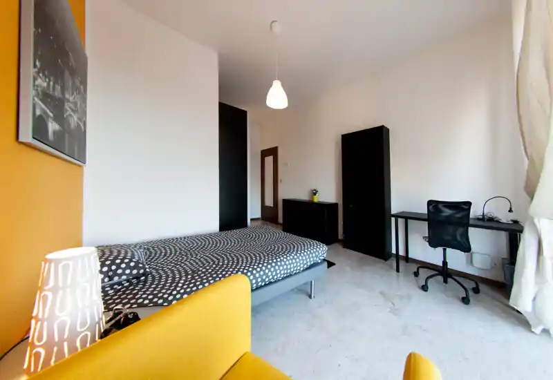 Bright, modern bedroom with a double bed, patterned bedding, a desk and storage units; warm yellow accents and natural light create a welcoming atmosphere.