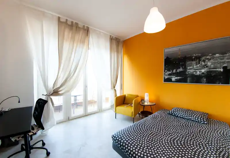 Bright, modern bedroom with a large window/door to a balcony, an accent orange wall, comfortable double bed with patterned bedding and a cozy armchair — ideal for a stylish city stay.
