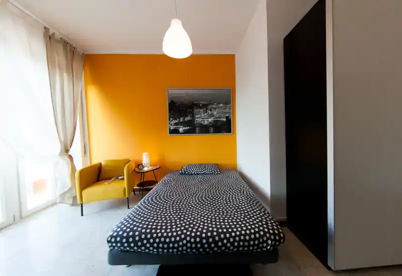 Bright, modern bedroom with a single bed facing the camera, a mustard accent wall and a comfortable armchair by the window. Large window drapes provide abundant natural light and contemporary decor adds warmth.