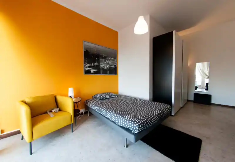 Bright, modern bedroom with a patterned double bed, mustard accent wall and a comfortable yellow armchair. Clean minimal styling and generous floor space make this room ideal for a stylish rental.
