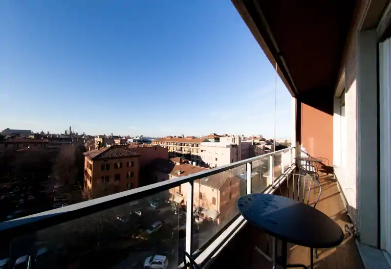 Balcone soleggiato con parapetto in vetro e ampia vista sulla città; piccolo tavolo e sedie per rilassarsi e godersi il panorama.