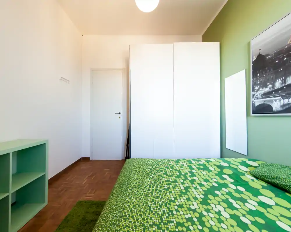Camera da letto luminosa e ben allestita con copriletto verde a motivi, armadio bianco e mensole semplici; linee pulite e colori coordinati creano un ambiente fresco e accogliente.