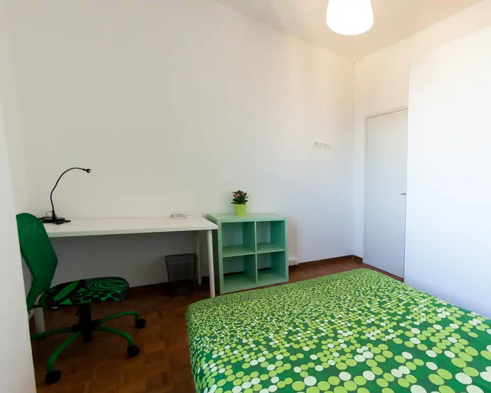 Camera da letto luminosa e ordinata con letto matrimoniale con copriletto verde a pois, una scrivania bianca e una sedia verde coordinata. Ambiente minimal e ben illuminato, ideale per annunci di affitto.