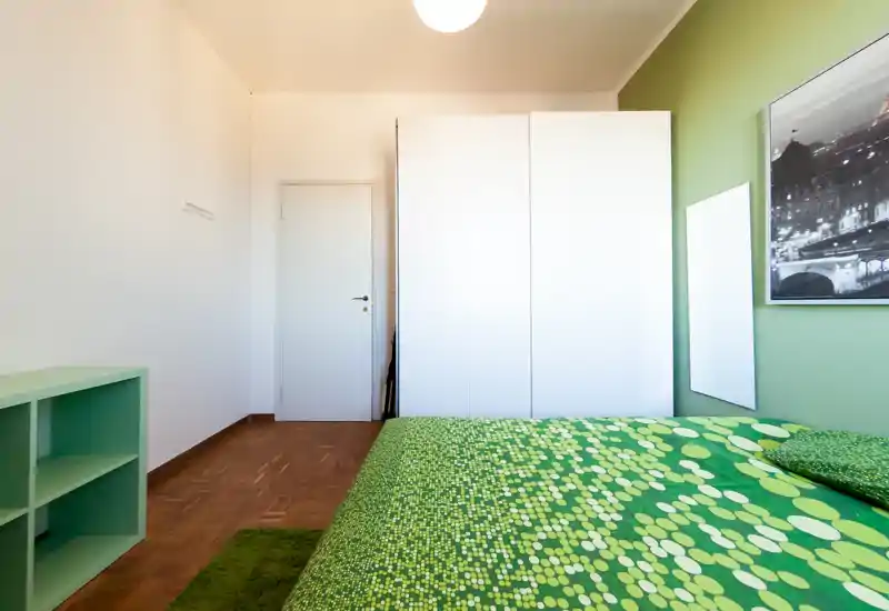 Camera da letto luminosa e ben allestita con copriletto verde a motivi, armadio bianco e mensole semplici; linee pulite e colori coordinati creano un ambiente fresco e accogliente.
