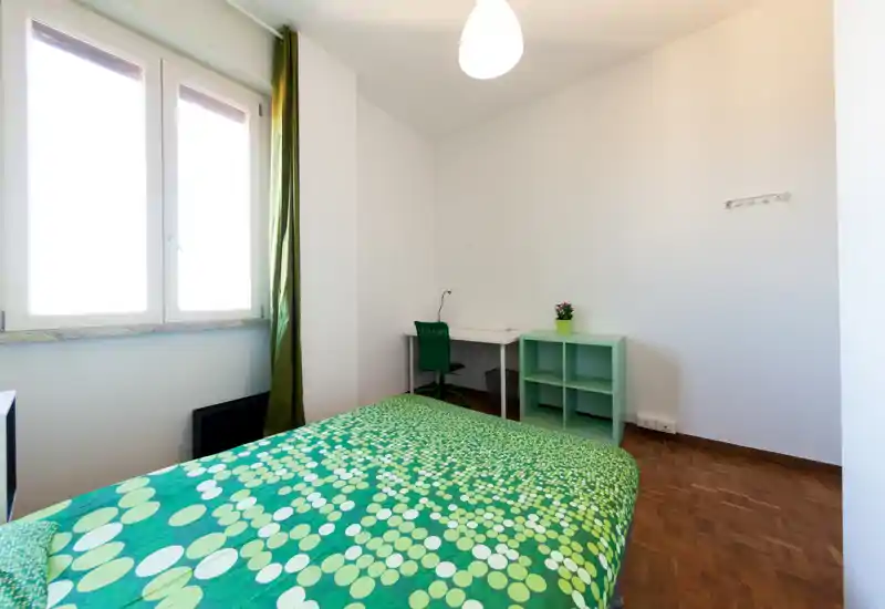 Camera da letto luminosa e semplice con letto matrimoniale a fantasia verde, scrivania e scaffalatura; luce naturale da una grande finestra che crea un'atmosfera ariosa.