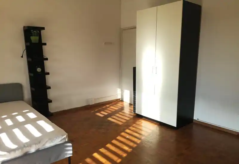 Dormitorio iluminado por el sol con una cama sencilla, suelo de parquet y un armario bicolor; la luz natural crea atractivos juegos de sombras.