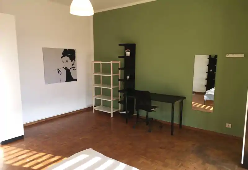 Rincón de dormitorio luminoso y minimalista con el borde del colchón visible, suelo de parquet, escritorio y estanterías junto a una pared verde.