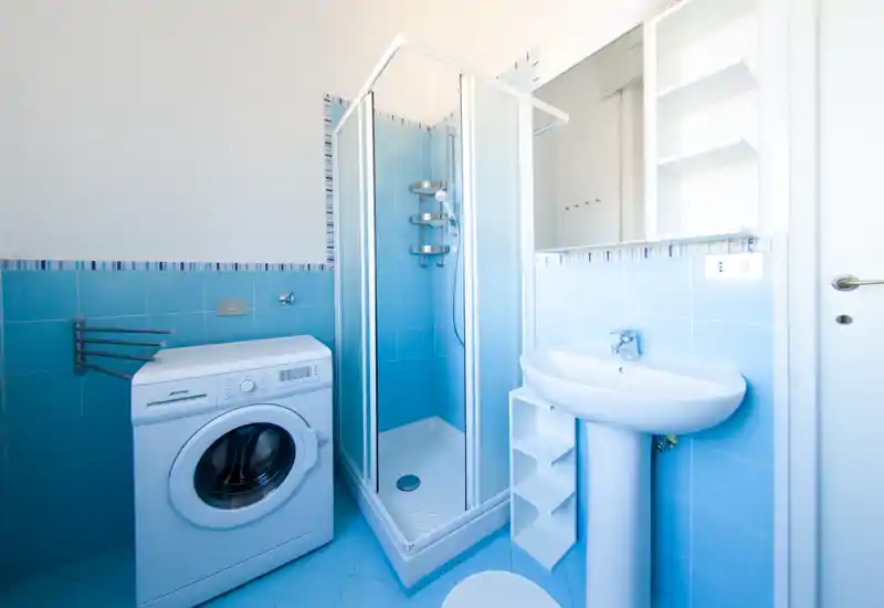 Bagno luminoso e ordinato con cabina doccia angolare, lavabo a colonna e lavatrice; piastrelle azzurre e mensole pratiche offrono funzionalità per bagno e lavanderia.