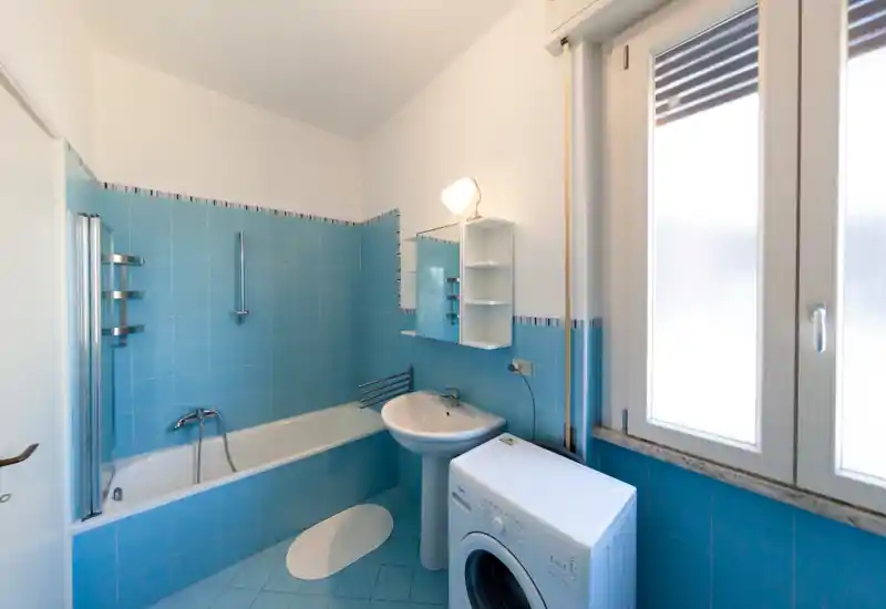 Bagno pulito e ben illuminato con vasca, lavabo a colonna, specchiera con armadietto e lavatrice accanto alla finestra. Piastrelle blu a parete e pavimento per un effetto fresco e uniforme.