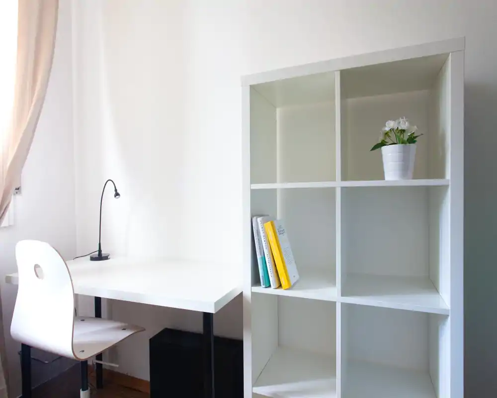 Rincón de trabajo luminoso y minimalista con escritorio blanco, silla moderna, lámpara pequeña y estantería blanca con libros y planta en maceta.