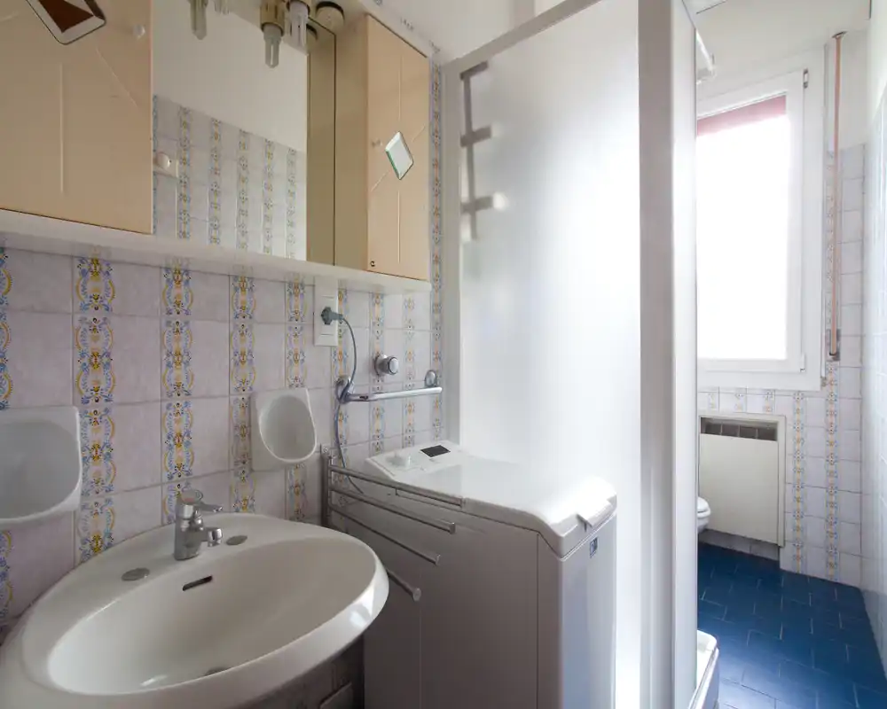 Baño compacto con lavabo, armario espejo, mampara de ducha translúcida y lavadora; la luz natural de la ventana ilumina el espacio con azulejos.