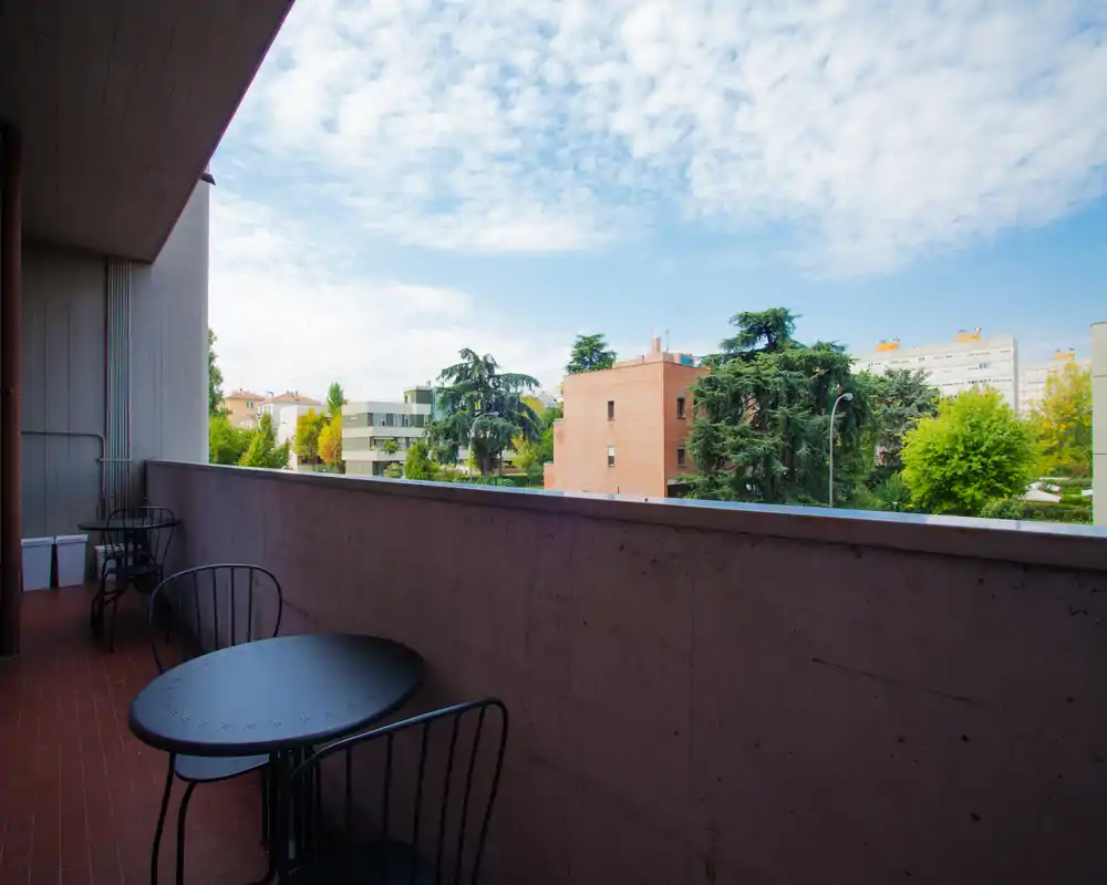 Balcón soleado con una pequeña mesa redonda y sillas de metal con vistas a árboles y edificios cercanos — espacio exterior luminoso y tranquilo.
