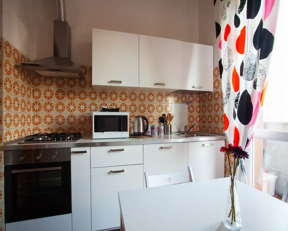 Cocina compacta y luminosa con azulejos de estilo retro, armarios blancos, encimera con fuego y horno. Mesa junto a la ventana con cortinas coloridas y un jarrón de flores que aportan calidez.