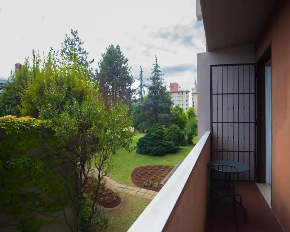 Pequeño balcón privado con mesa y sillas con vistas a un jardín cuidado y árboles maduros — ideal para tomar un café por la mañana y disfrutar de momentos tranquilos al aire libre.