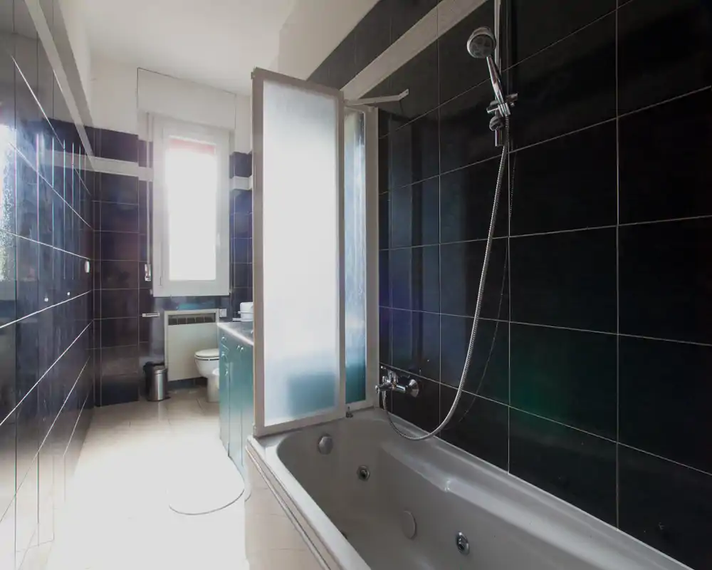 Baño moderno con bañera y ducha de pared, paredes azulejadas oscuras y mampara de vidrio esmerilado junto a la bañera; ventana que aporta luz natural.