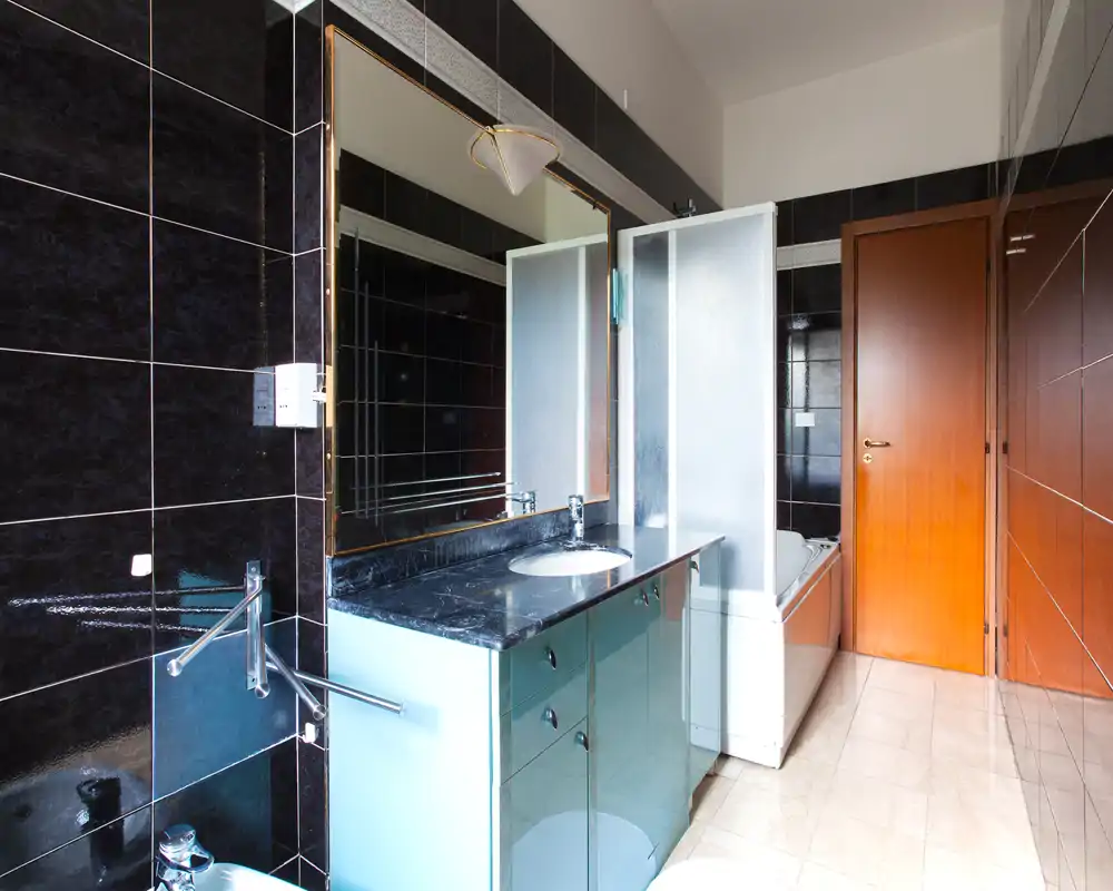 Baño moderno y limpio con azulejos negros brillantes, lavabo sobre encimera oscura y bañera/ducha con mampara de vidrio esmerilado.