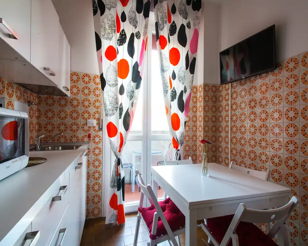 Cocina compacta con una pequeña mesa junto a la puerta del balcón que aporta mucha luz. Azulejos decorativos y cortinas llamativas dan personalidad; los muebles blancos mantienen el aspecto limpio.