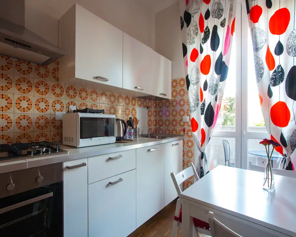 Cocina compacta y luminosa con armarios blancos, azulejos retro estampados, placa de gas, microondas y una pequeña mesa junto a la puerta que da al balcón.