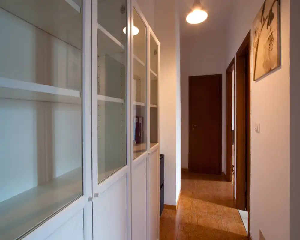 Entrada/pasillo luminoso y estrecho con armarios blancos empotrados a la izquierda, suelo de baldosas cálidas y puerta de madera al fondo, ideal para mostrar almacenamiento de entrada y el paso.
