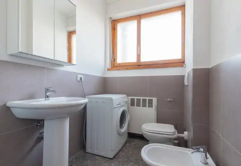 Baño compacto y funcional con lavabo pedestal, bidé, inodoro y una lavadora bajo una ventana luminosa con marco de madera.