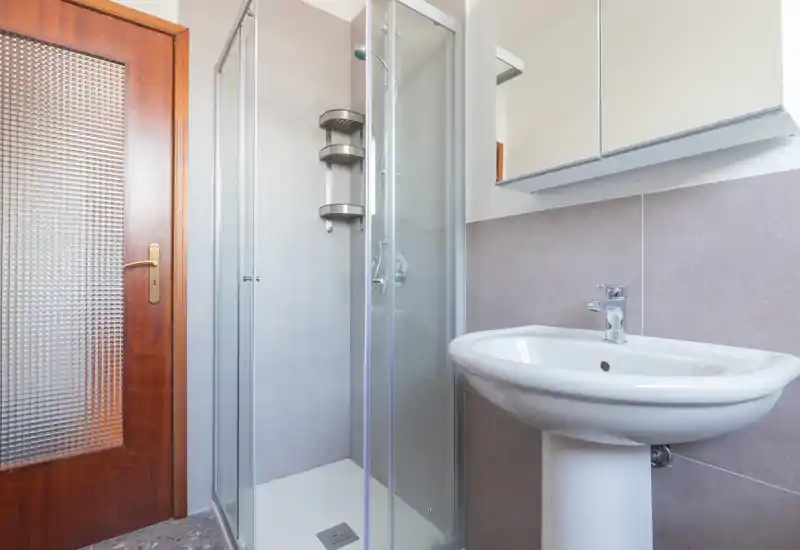 Baño limpio y compacto con ducha con mampara de cristal, lavabo de pedestal, revestimiento cerámico y armario espejo.