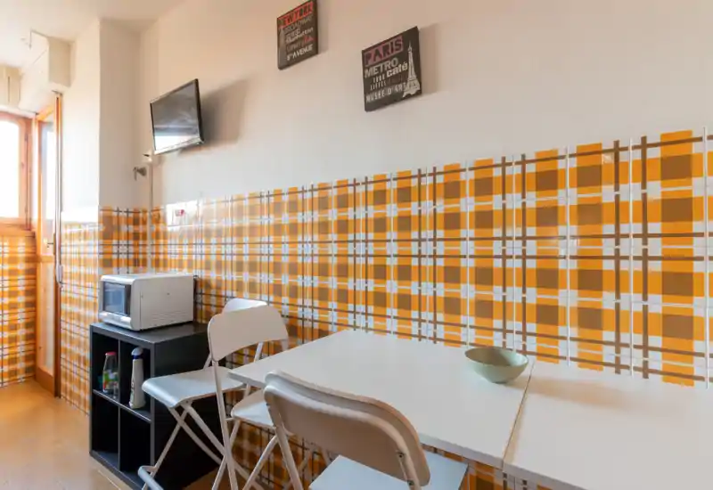 Cocina pequeña y luminosa con azulejos naranjas estampados, mesa blanca y sillas plegables, microondas y TV en la pared—ideal para estancias cortas.