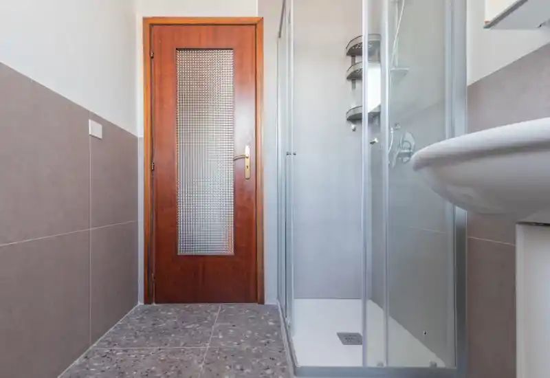 Baño compacto y limpio con mampara de ducha de vidrio, lavabo blanco y puerta de madera; luminoso y bien alicatado.