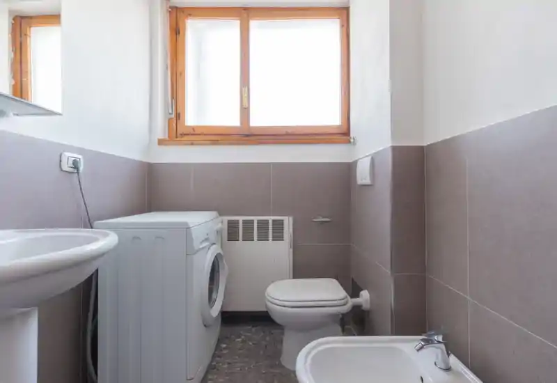 Baño compacto y bien iluminado con lavabo, bidé, inodoro y una lavadora bajo una ventana de marco de madera.