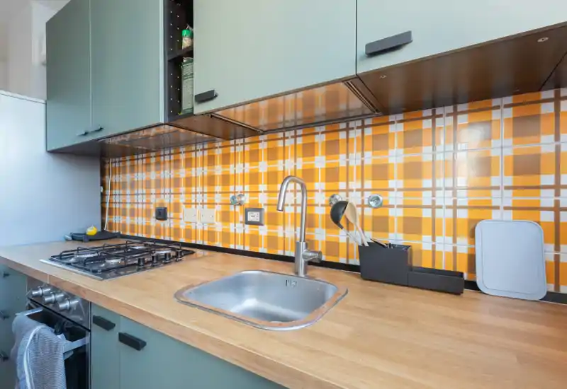 Cocina compacta y contemporánea con encimera de madera, fregadero de acero, placa de gas y un llamativo salpicadero a cuadros naranja que aporta carácter y calidez.