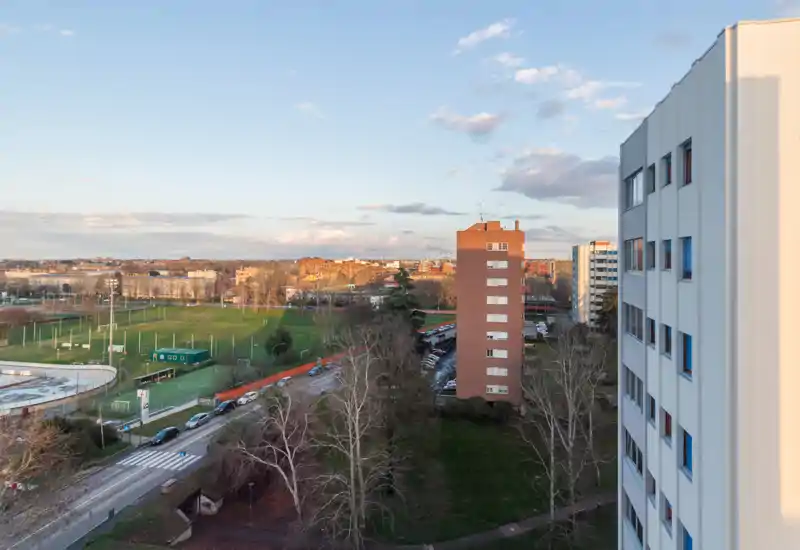 Amplia vista exterior desde un piso alto que muestra un parque con campos deportivos, edificios residenciales y una carretera; la luz del atardecer crea una atmósfera tranquila — ideal para destacar la vista del balcón.