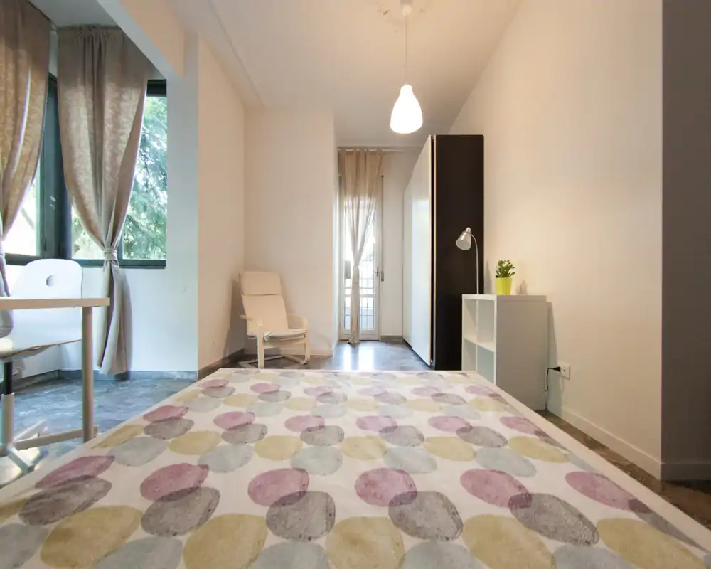 Chambre lumineuse et moderne avec housse de couette à motifs, fauteuil de lecture et grandes fenêtres avec rideaux laissant passer la lumière naturelle. Mobilier minimal et bien rangé avec petite étagère et armoire, idéale pour un séjour confortable.