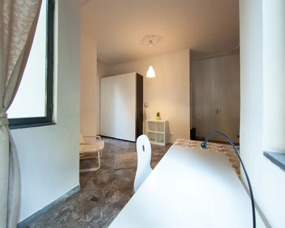 Coin chambre/studio compact avec bureau près de la fenêtre, armoire, fauteuil et éclairage doux ; espace lumineux et fonctionnel adapté à une personne seule.
