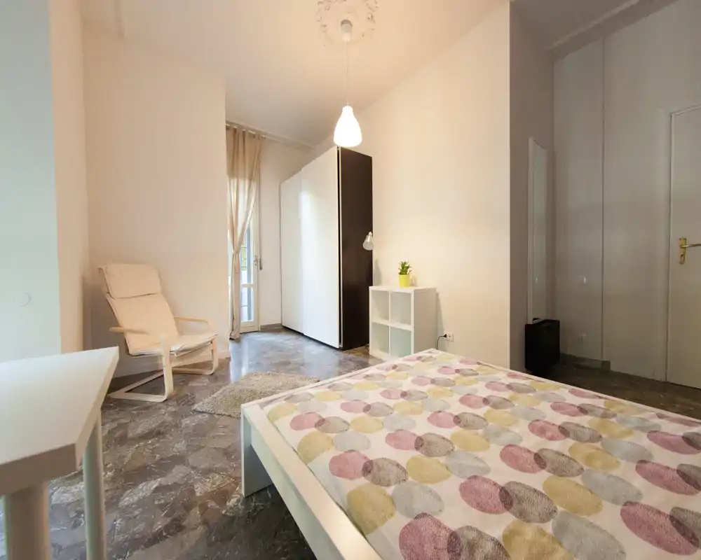 Chambre moderne et lumineuse avec couette à motifs, cadre de lit blanc et meubles minimalistes; lumière naturelle douce et un fauteuil confortable pour une ambiance chaleureuse.