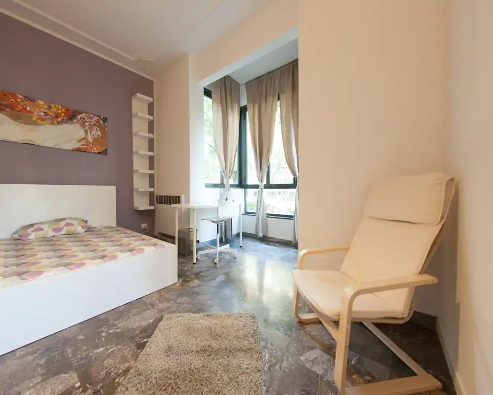 Chambre moderne et lumineuse avec lit blanc, couette à motifs, tapis douillet et fauteuil près de la fenêtre pour un coin lecture agréable.