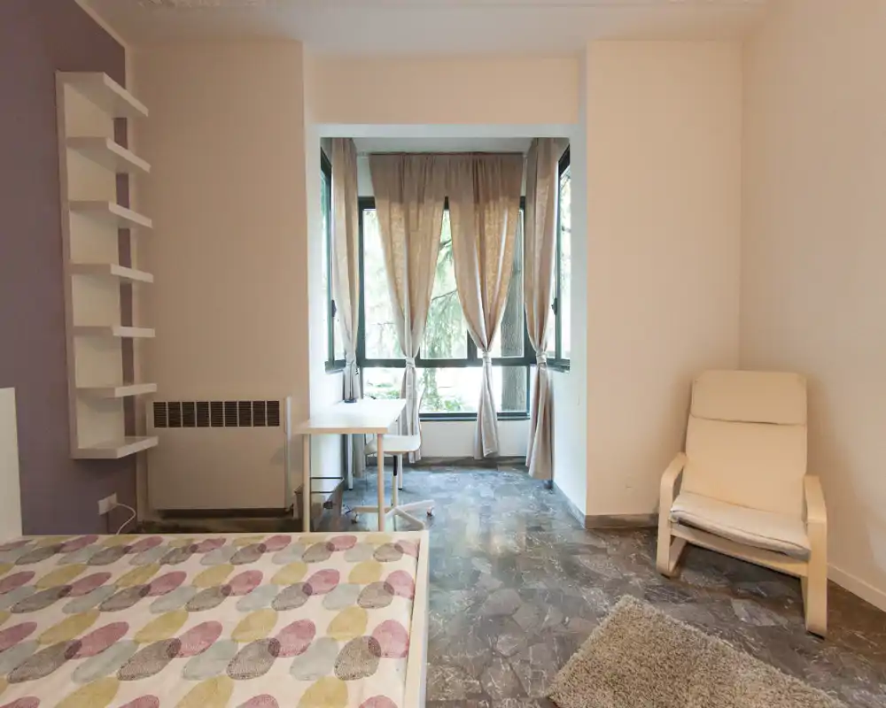 Chambre lumineuse avec lit à motif au premier plan, un fauteuil confortable et un petit bureau près de grandes fenêtres apportant beaucoup de lumière naturelle.