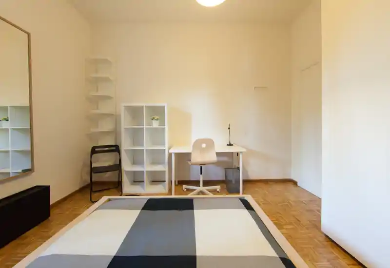 Minimalist bedroom with a double bed in the foreground, simple desk and shelving against a neutral wall, and warm parquet flooring. Bright, uncluttered space suitable for rental photos.