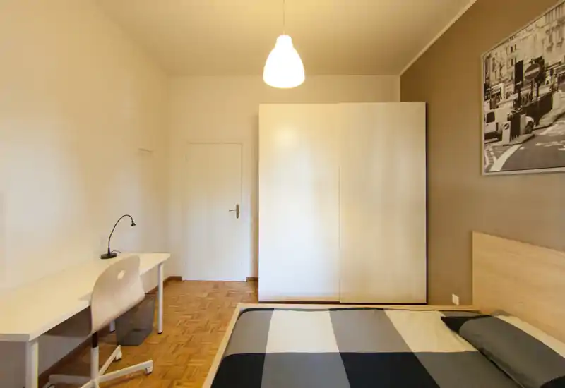 Comfortable, well-lit bedroom with a double bed, a large white wardrobe and a work desk. Neutral tones, parquet flooring and framed wall art create a calm, modern atmosphere ideal for short-term rentals.