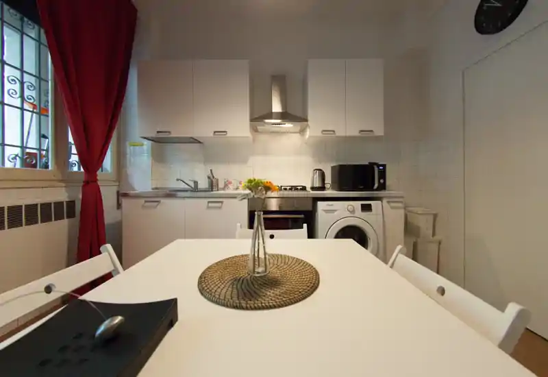 Bright compact kitchen with a dining table in the foreground, white cabinets, gas stove, microwave and washing machine; decorative vase on the table and red curtains at the window.