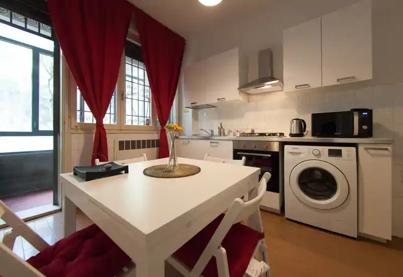 Bright, well-equipped kitchen with a white dining table, four chairs, stove, oven, washing machine and microwave. Red curtains and a small centerpiece add a warm, homely touch.