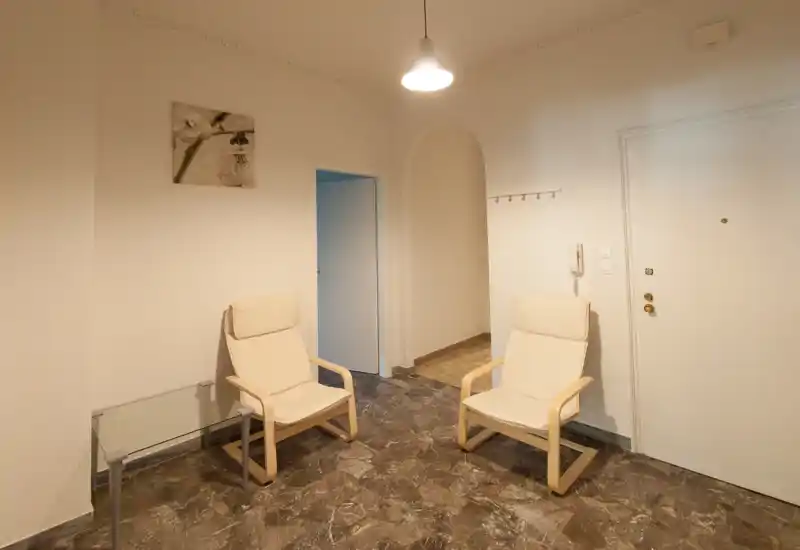 Small, simply furnished living area with two cream armchairs, pendant lighting and marble-effect floor; neutral walls and minimal décor create a calm, functional sitting space.