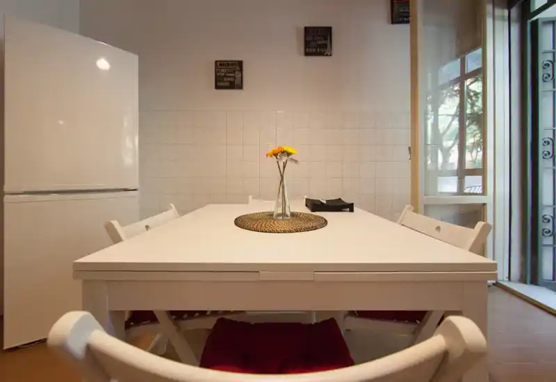 Bright, well-composed dining area with a white table, chairs and a vase centerpiece; natural light from a large glass door creates an inviting atmosphere.